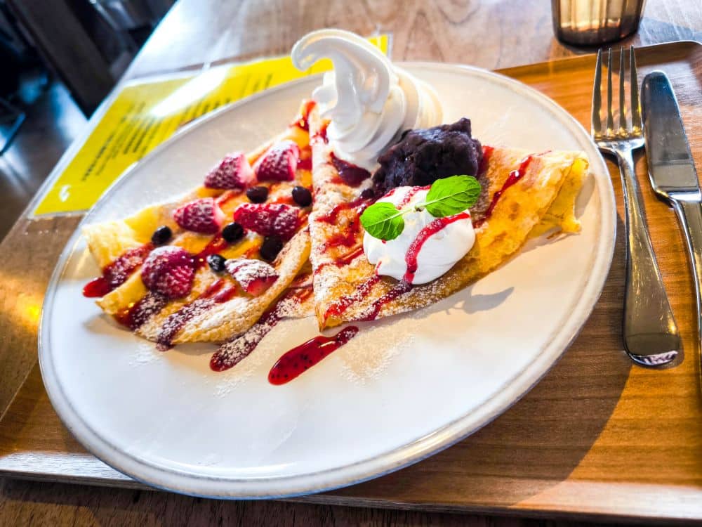 Auf einem Holztablett mit Gabel und Messer liegt ein Teller mit zwei Fruchtscheiben, Schlagsahne, Beerensoße, einem Strudel Softeis und Minzblättern - und bringt Ihnen die Süße des Enoshima-Meeres an den Tisch.