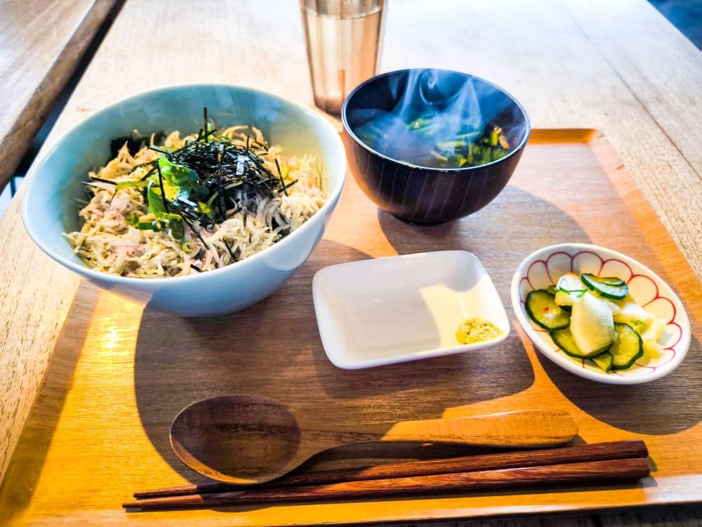 Eine japanische Mahlzeit auf einem Holztablett enthält Reis nach Enoshima-Art mit geraspeltem Seetang, eine Schale mit gedämpfter Suppe, eingelegte Gurken, Dip-Sauce, einen Löffel und Stäbchen.