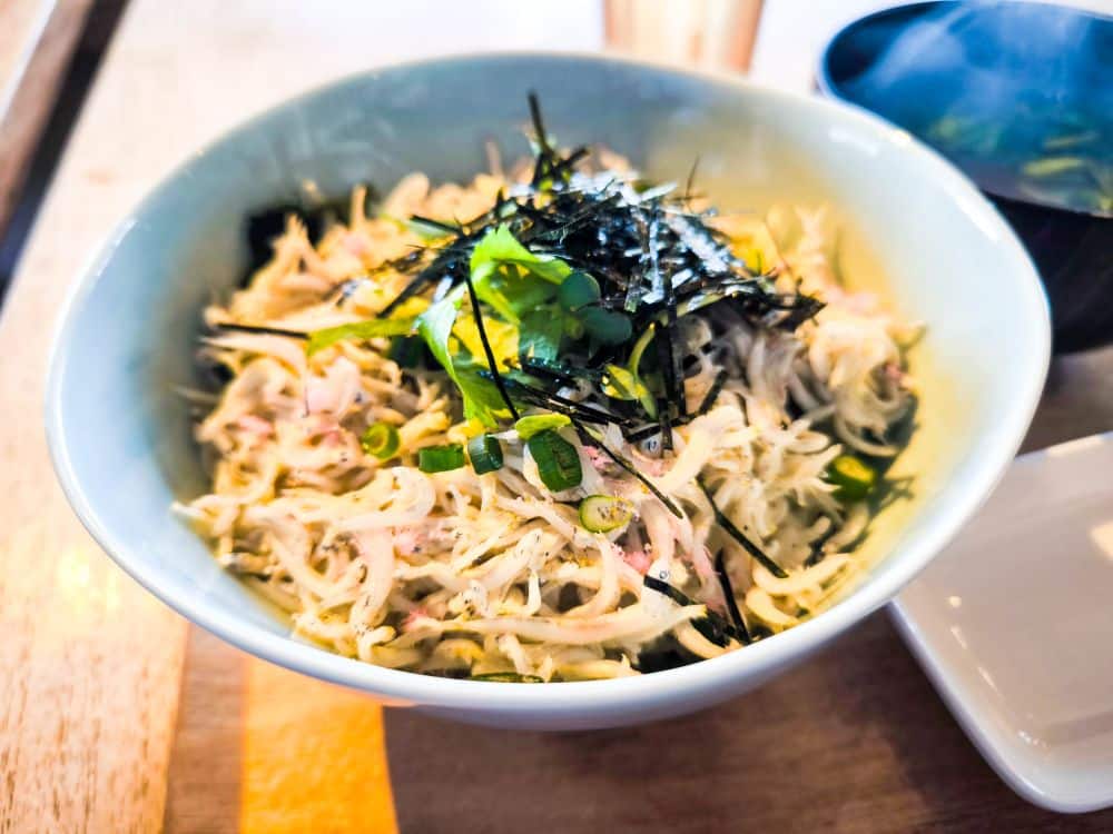 Eine Schüssel mit zerkleinertem Hühnersalat, garniert mit geschnittenen grünen Zwiebeln und dünnen Nori-Seetangstreifen, inspiriert von den Aromen aus Enoshima, serviert auf einem Holztisch mit einer kleinen Schale Suppe im Hintergrund.