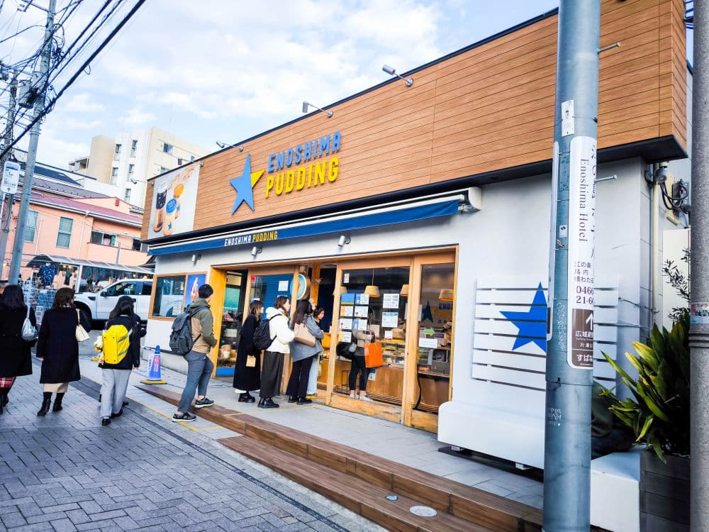 Die Leute stehen in der Schlange vor Enoshima Pudding, einem modernen Geschäft mit blauem Stern-Logo, Holzvertäfelung und großen Fenstern an der belebten Straße von Enoshima, die von Gebäuden und Stromleitungen gesäumt ist, unter einem klaren Himmel.