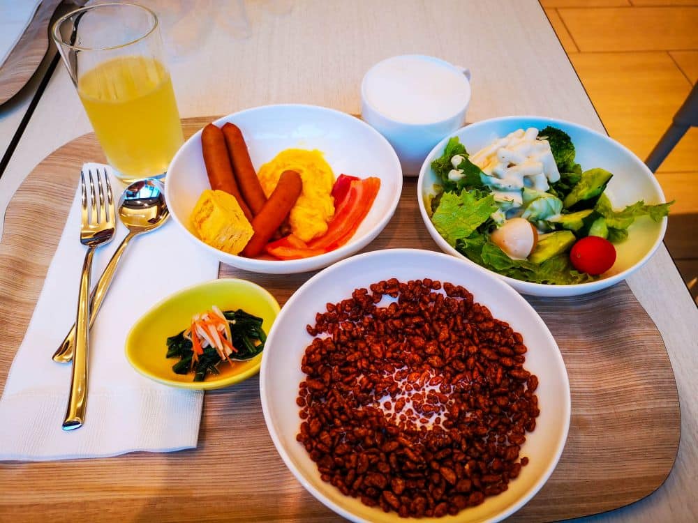 Frühstückstablett im Tobu mit einer Schale Schokomüsli und Milch, einem Teller mit Rührei, Würstchen und Gemüse, einem frischen Salat mit Dressing, einem kleinen Teller mit Grünzeug, einem Glas Saft und einer Tasse Milch.