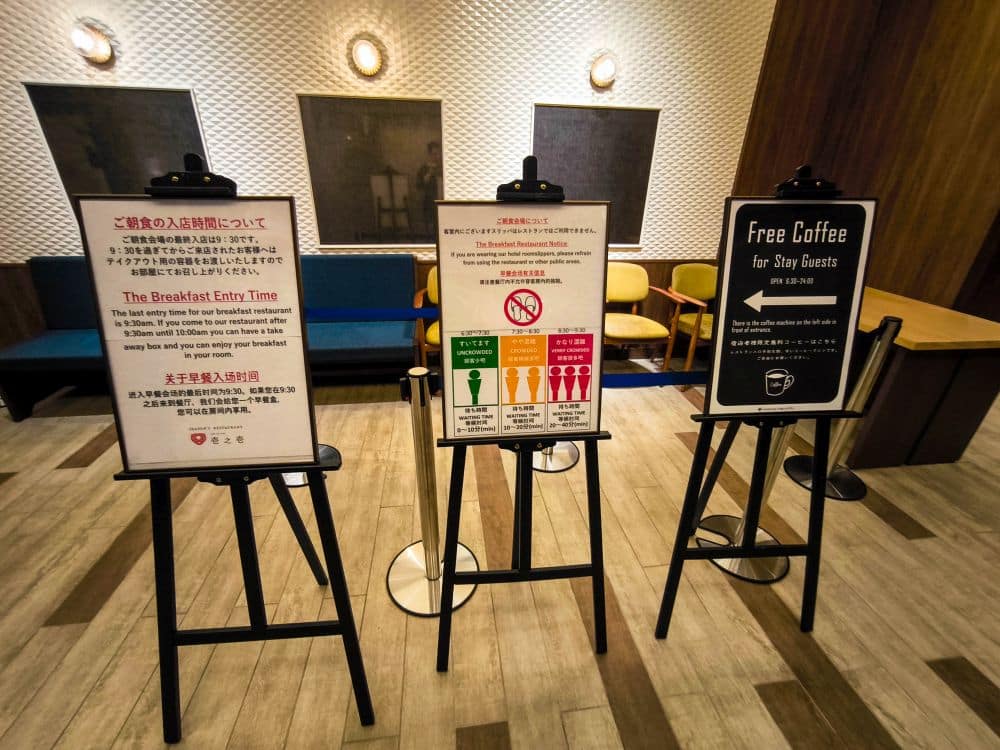 In der Lobby des Tobu-Hotels stehen drei Schilder auf Staffeleien, auf denen die Einlasszeiten für das Frühstück, die Regeln und ein Hinweis auf kostenlosen Kaffee für die Gäste stehen. Dahinter stehen moderne Stühle, ein kleiner Tisch und gemusterte Wanddekoration.