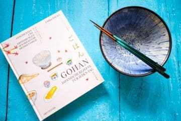 Ein japanisches Kochbuch, eine blaue Keramikschüssel und ein Paar Essstäbchen ruhen auf einer hellblauen Holzfläche. Der Einband des Kochbuchs zeigt Illustrationen von Lebensmitteln und einen japanischen und deutschen Text, der die Geschichte von Gohan wiedergibt.