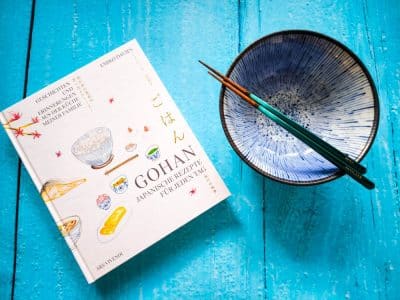 Ein japanisches Kochbuch, eine blaue Keramikschüssel und ein Paar Essstäbchen ruhen auf einer hellblauen Holzfläche. Der Einband des Kochbuchs zeigt Illustrationen von Lebensmitteln und einen japanischen und deutschen Text, der die Geschichte von Gohan wiedergibt.