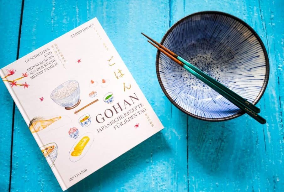 Ein japanisches Kochbuch, eine blaue Keramikschüssel und ein Paar Essstäbchen ruhen auf einer hellblauen Holzfläche. Der Einband des Kochbuchs zeigt Illustrationen von Lebensmitteln und einen japanischen und deutschen Text, der die Geschichte von Gohan wiedergibt.