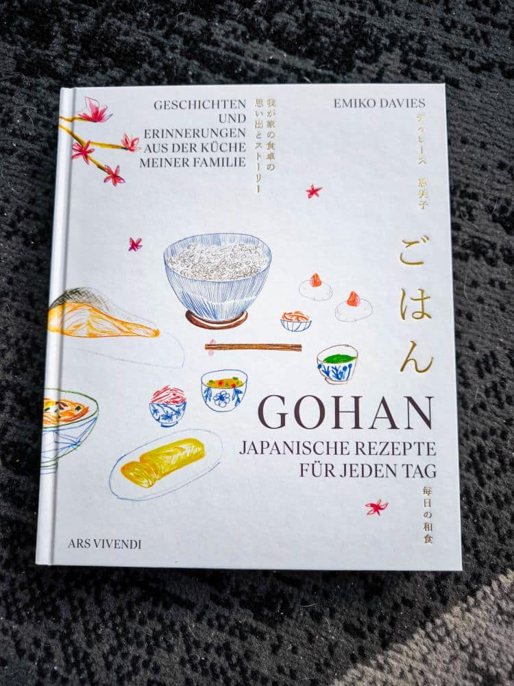Ein Kochbuch mit dem Titel Gohan: Japanische Rezepte für jeden Tag von Emiko Davies, mit handgezeichneten Illustrationen japanischer Küche auf dem Cover, liegt auf einer dunklen, strukturierten Oberfläche.