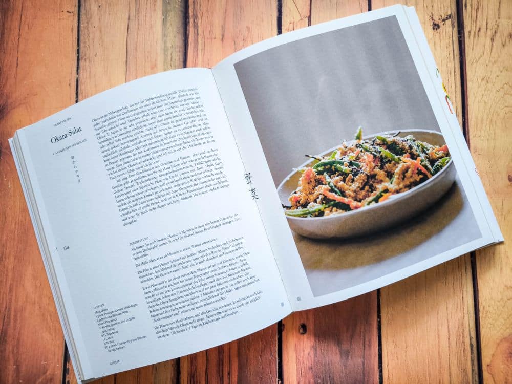 Ein offenes Kochbuch liegt auf einer Holzoberfläche und zeigt links eine Geschichte über Okra-Salat und rechts eine große Schale mit farbenfrohem Okra-Salat.