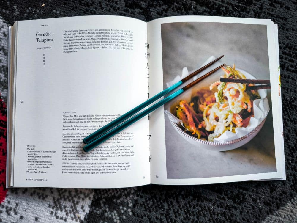 Ein aufgeschlagenes Kochbuch mit einem Gemüsetempura-Rezept auf der linken Seite und einem Foto des Gerichts auf der rechten Seite, mit einem Paar blauer Essstäbchen, die quer über die Seiten gelegt sind, hält Gohans kulinarische Geschichte fest.
