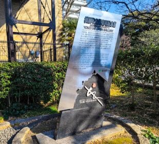 Ein reflektierendes Metallschild mit japanischem und englischem Text steht auf einer kleinen begrünten Fläche. Es kennzeichnet den Standort der Domänenglocke von Yokohama und enthält eine Karte und ein Logo, das auf die Geschichte des Gebiets und seine Verbindung zum großen Kanto-Earthquake verweist.