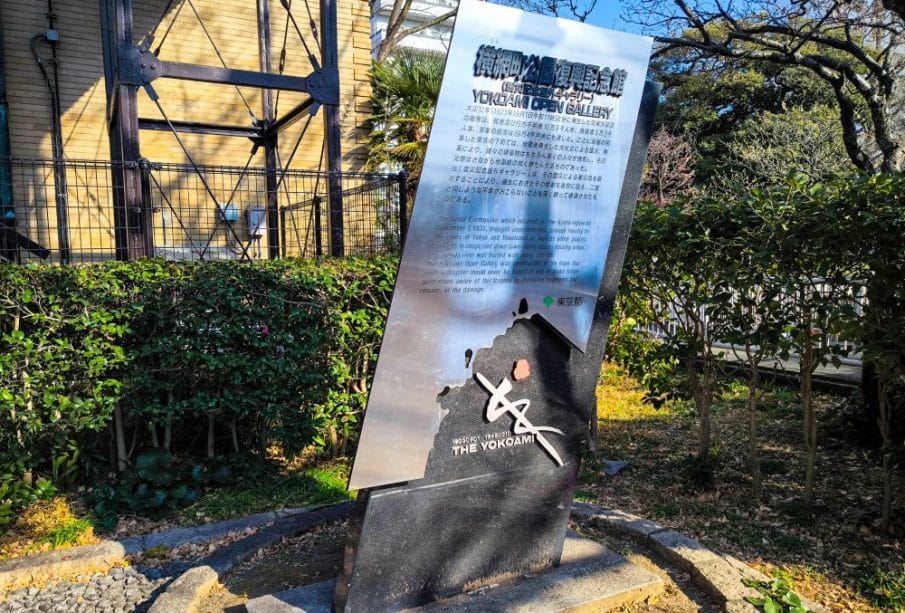 Ein reflektierendes Metallschild mit japanischem und englischem Text steht auf einer kleinen begrünten Fläche. Es kennzeichnet den Standort der Domänenglocke von Yokohama und enthält eine Karte und ein Logo, das auf die Geschichte des Gebiets und seine Verbindung zum großen Kanto-Earthquake verweist.