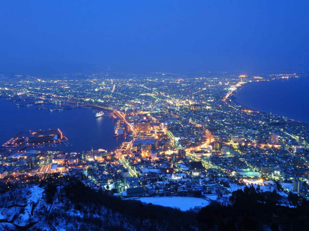 Luftaufnahme einer Küstenstadt in Hokkaido in der Abenddämmerung. Die Lichter der Stadt beleuchten Straßen und Gebäude, eine geschwungene Küstenlinie, einen Hafen mit Docks und schneebedeckte Hügel im Vordergrund.