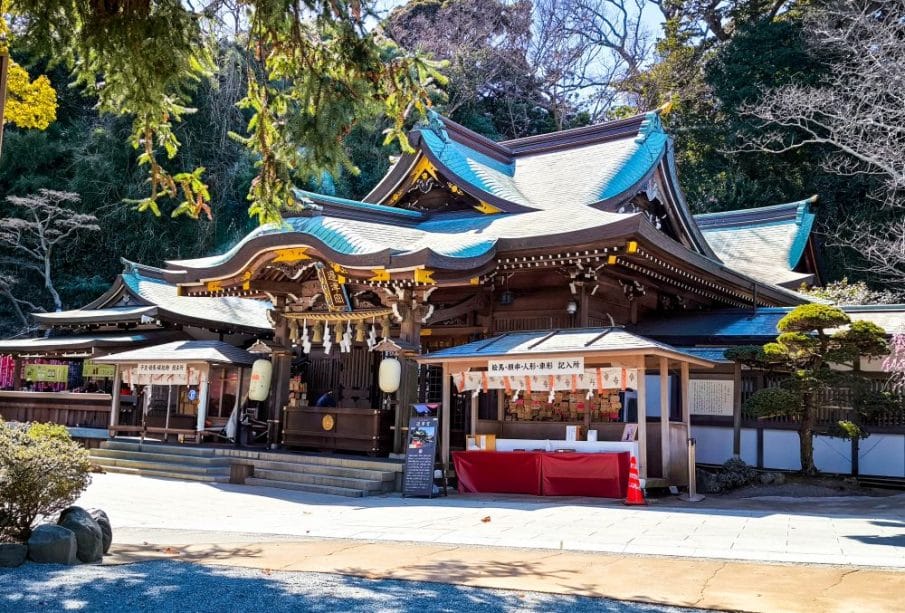 Der Hetsunomiya, ein traditioneller japanischer Shinto-Schrein mit verschnörkelter Holzarchitektur und geschwungenen blauen Ziegeldächern, ist von Bäumen umgeben; vor dem Schrein stehen eine rote Tafel und kleine Verkaufsstände.