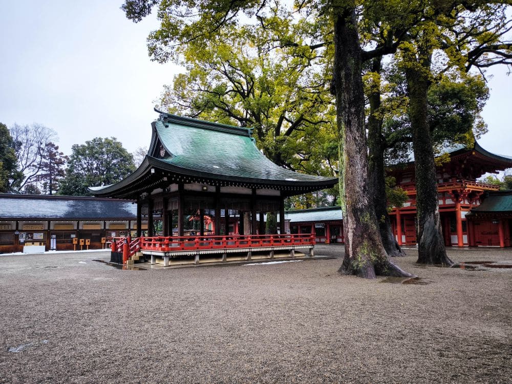 Ein traditioneller Hikawa-Schrein mit grünem Ziegeldach, rotem Geländer und umstehenden Bäumen steht in einem ruhigen, offenen Hof mit Kiesboden.