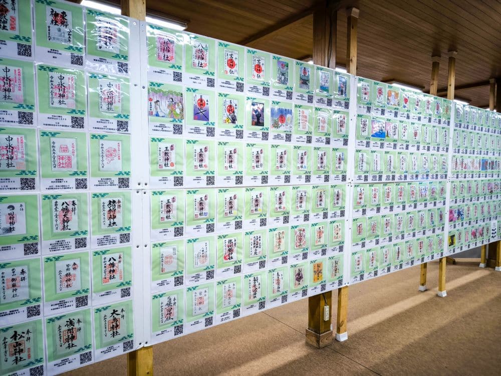 Eine große Schautafel in Hikawa, die mit Reihen bunter Karten mit japanischer Kalligrafie und Kunstwerken bedeckt ist, ist in einem Holzgebäude angebracht. Die übersichtlich angeordneten Karten enthalten auch QR-Codes, die den Zugang zu weiteren Informationen erleichtern.