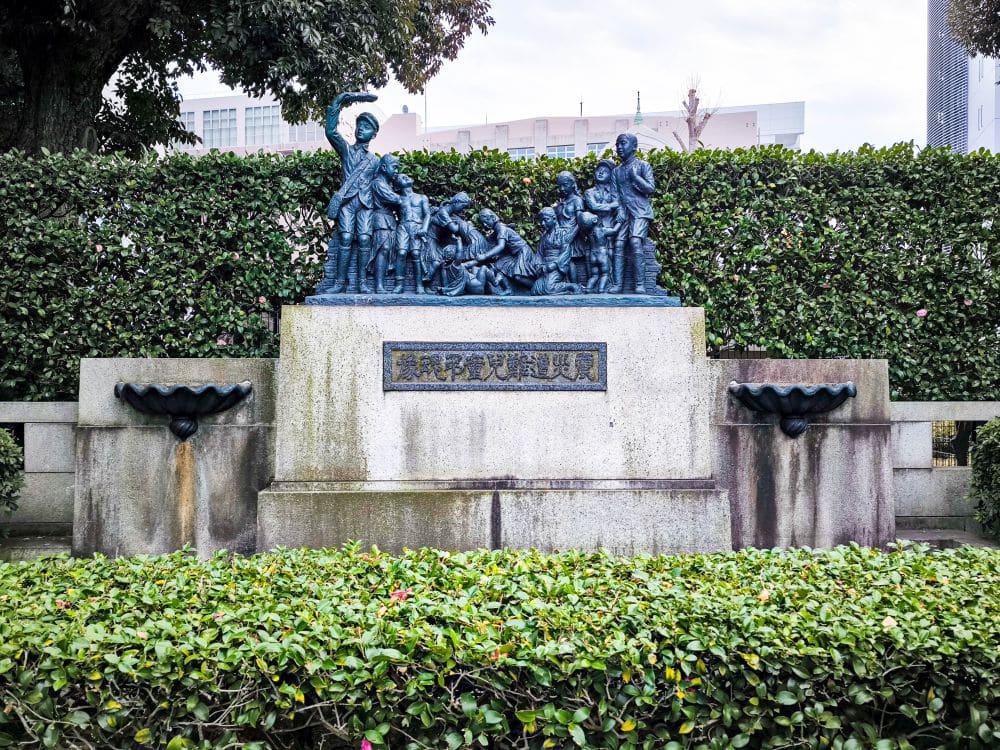 Eine Bronzestatue mit mehreren Kindern und einem Erwachsenen steht auf einem Steinsockel mit einer Gedenktafel, umgeben von Grünpflanzen. Diese Außenanlage in der Nähe des Gedenkmuseums erinnert an die Opfer des großen Kanto-Erdbebens.