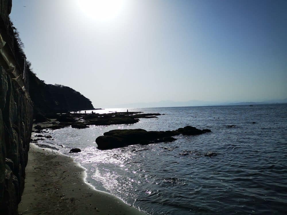 Eine felsige Enoshima-Küste bei Sonnenuntergang mit sanften Wellen, silhouettierten Klippen und einer hellen Sonne am klaren Himmel, die schimmernde Reflexe auf dem Wasser erzeugt. In der Ferne stehen ein paar Menschen auf den Felsen.