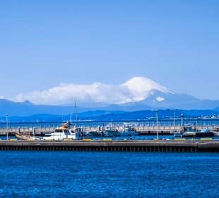 Ein malerischer Blick auf den Hafen von Enoshima mit angedockten Booten und tiefblauem Wasser, vor dem Hintergrund des schneebedeckten Gipfels des Mount Fuji, der sich unter einem klaren, strahlend blauen Himmel erhebt. Start