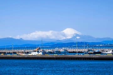Ein malerischer Blick auf den Hafen von Enoshima mit angedockten Booten und tiefblauem Wasser, vor dem Hintergrund des schneebedeckten Gipfels des Mount Fuji, der sich unter einem klaren, strahlend blauen Himmel erhebt. Start