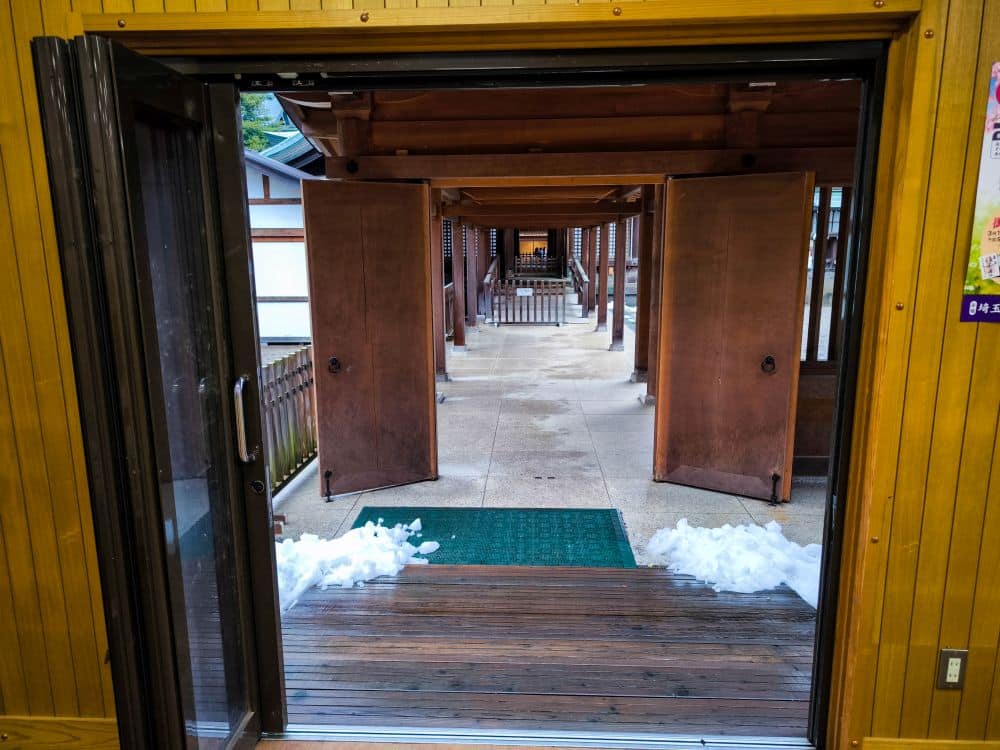 Blick durch eine offene Tür auf einen überdachten hölzernen Hikawa-Gang, der auf beiden Seiten mit offenen Türen gesäumt ist, mit Schneeflecken in der Nähe des Eingangs und Licht, das durch den Gang scheint.