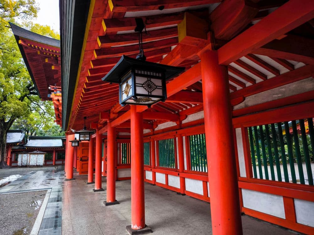 Überdachter Gang mit leuchtend roten Säulen und traditionellen Laternen an einem Hikawa-Schrein, mit einem Ziegeldach und grünen Bäumen im Hintergrund.