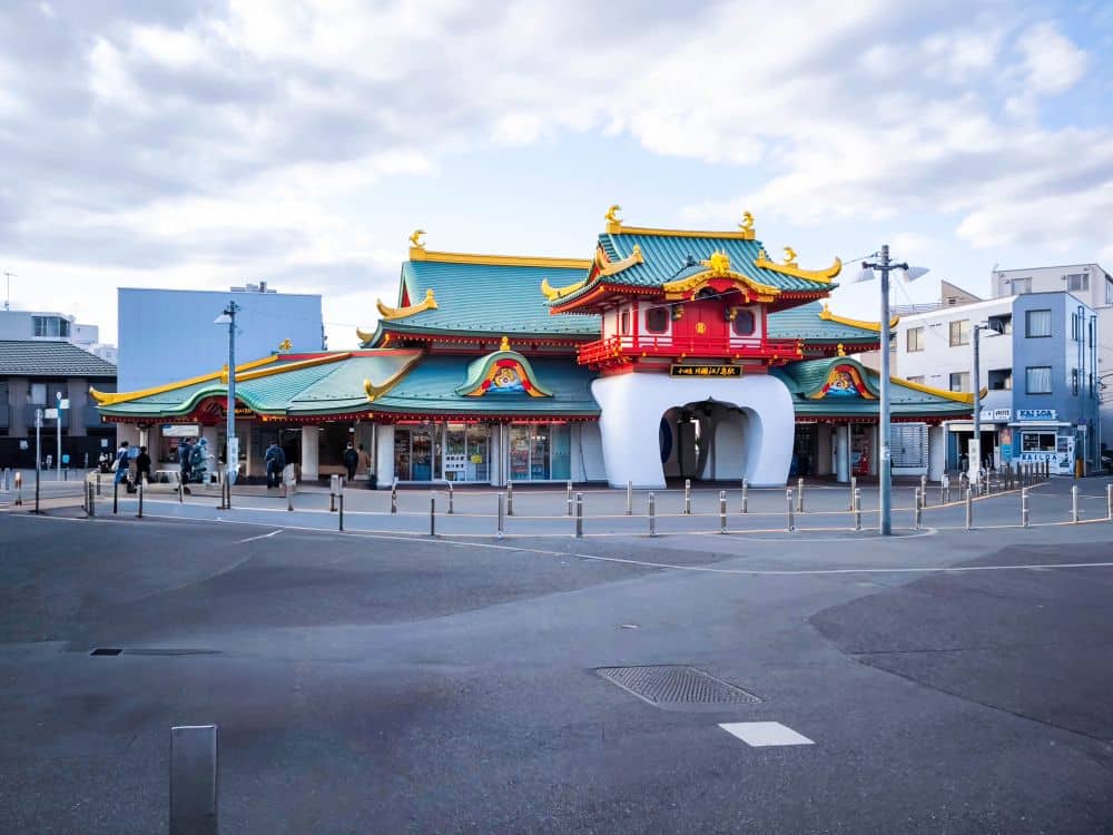 Ein farbenfrohes Gebäude mit verschnörkelten grünen, roten und goldenen Dächern steht unter einem teilweise bewölkten Himmel in Enoshima. In der Nähe gehen Menschen spazieren, und moderne Stadtgebäude umgeben das Gebäude.
