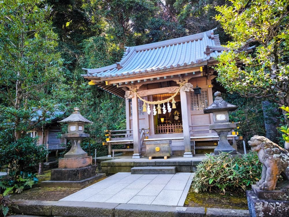Ein traditioneller japanischer Shinto-Schrein, bekannt als Hetsunomiya, mit Ziegeldach, Steinlaternen, Wächterstatue und umgeben von üppigen grünen Bäumen und Blättern.