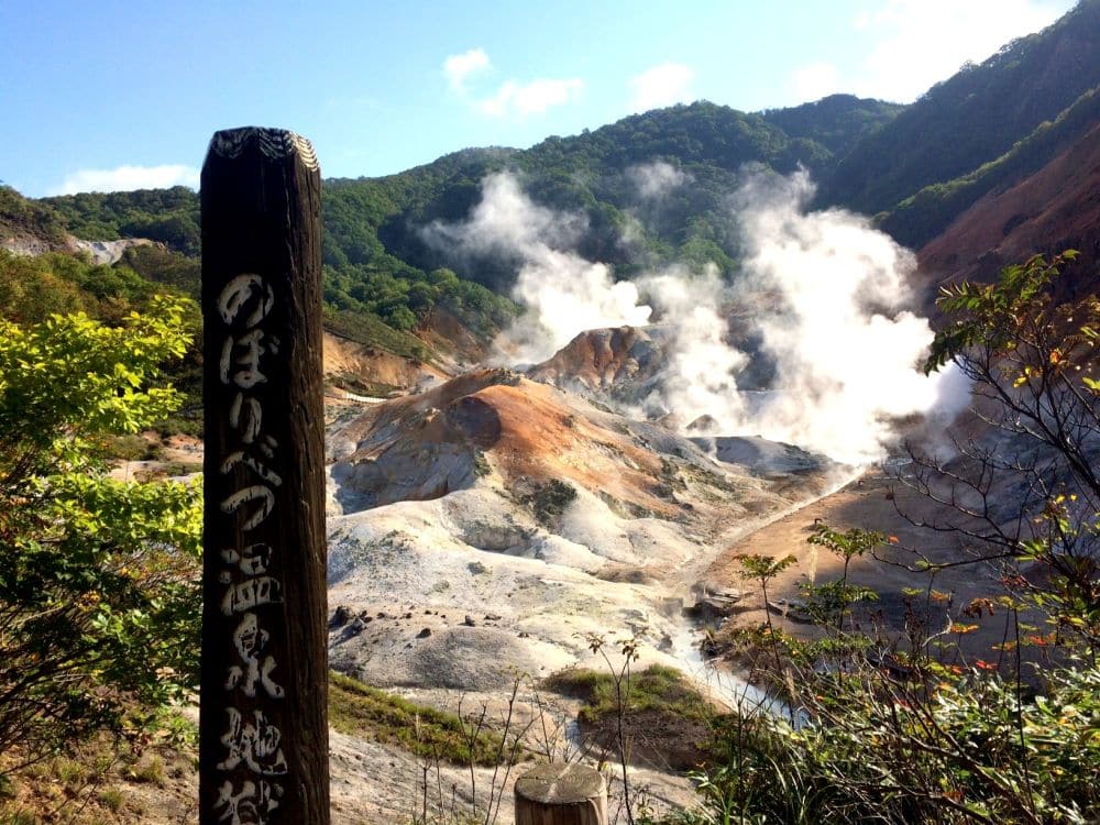 Ein dampfendes Vulkantal in Hokkaido mit zerklüftetem, farbenfrohem Terrain und aufsteigenden Dämpfen, umgeben von grünen Hügeln. Ein Holzpfosten mit japanischer Schrift steht im Vordergrund unter einem klaren, blauen Himmel.