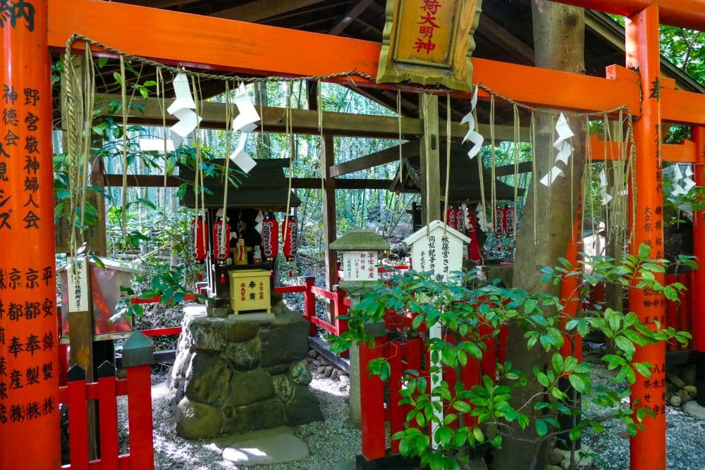 Ein kleiner Shinto-Schrein mit roten Torii-Toren, traditionellen Dekorationen und Talismanen aus Papier steht friedlich inmitten von Grünpflanzen und Bambus und erinnert an einen ruhigen japanischen Tempel im Freien.