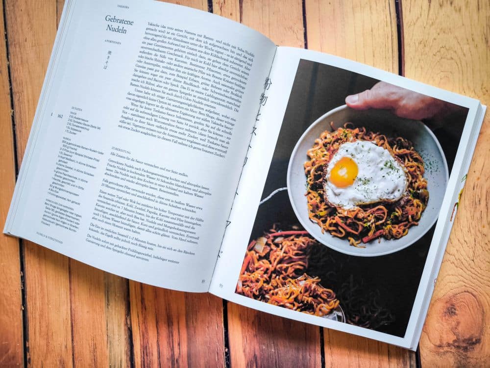 Ein aufgeschlagenes Kochbuch auf einer hölzernen Unterlage zeigt auf der linken Seite ein Rezept für gebratene Nudeln und auf der rechten Seite ein Foto von Nudeln mit Spiegelei in einer weißen Schüssel, die die einfache Geschichte der Hausmannskost einfangen.