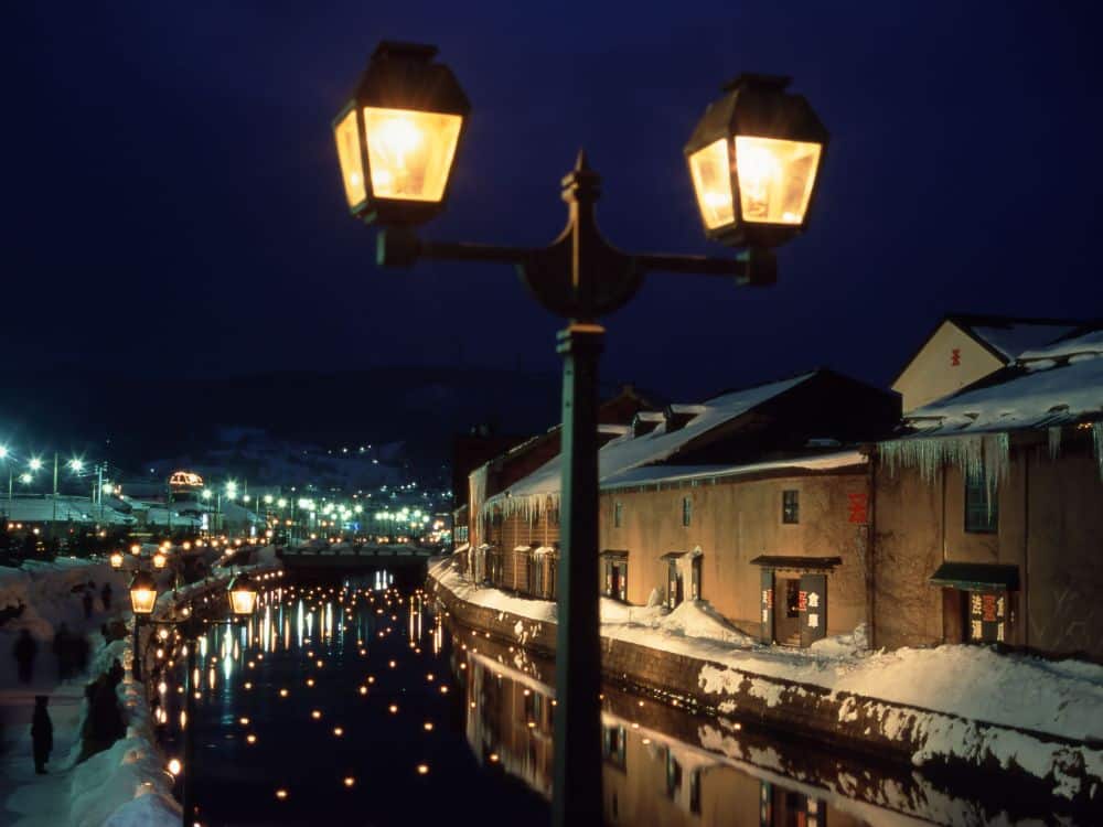 Ein verschneiter Kanal in Hokkaido bei Nacht wird von leuchtenden Straßenlaternen erhellt, deren Spiegelungen im Wasser schimmern. Traditionelle Gebäude säumen den Kanal, während in der Ferne die Lichter der Stadt unter einem dunkelblauen Himmel funkeln.