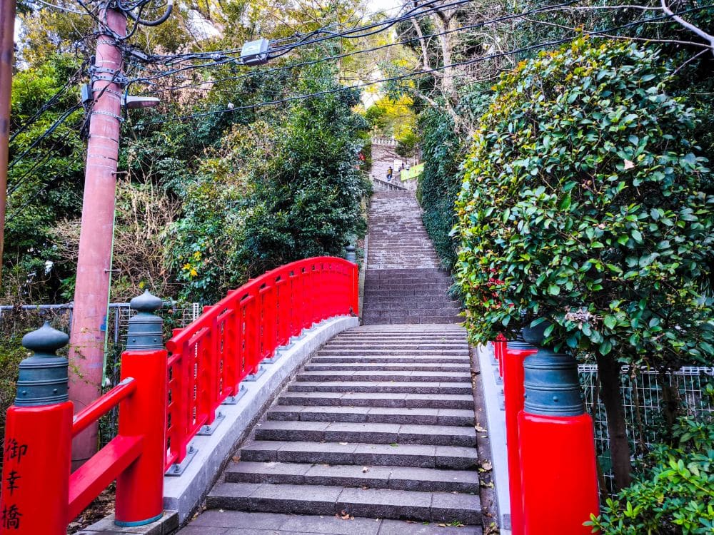 Eine leuchtend rote Bogenbrücke in Enoshima führt zu einer langen Steintreppe, umgeben von grünen Bäumen und Sträuchern, mit Stromleitungen und einem Holzmast auf der linken Seite. Die Szene wirkt ruhig und einladend.