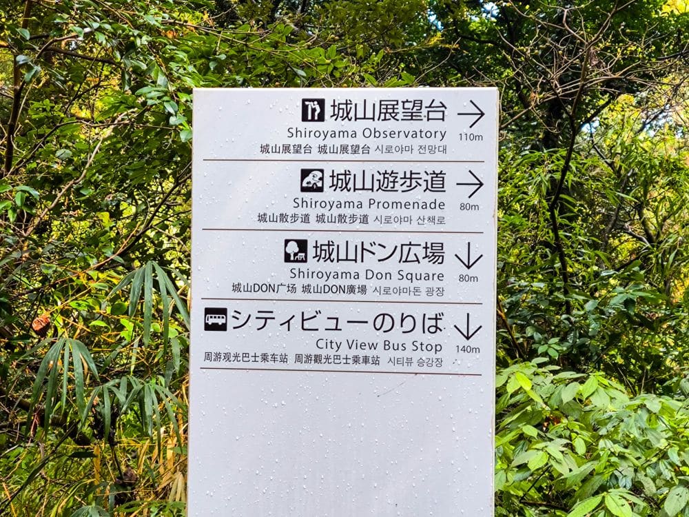 Wegweiser in Japanisch, Englisch und Koreanisch in einer üppigen Grünanlage in Kagoshima, die zur Aussichtsplattform des Shiroyama-Parks, zur Shiroyama-Promenade, zum Don-Platz und zur Bushaltestelle City View führen und einen Blick auf Sakurajima bieten - mit Entfernungsangaben.