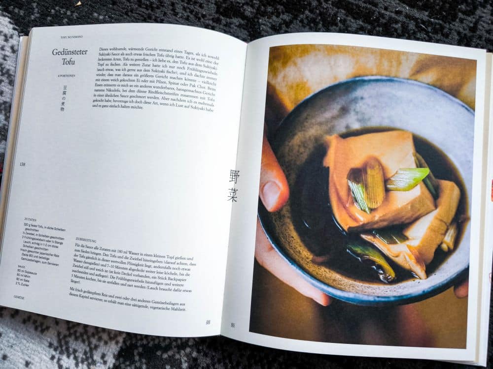 Ein aufgeschlagenes Kochbuch zeigt auf der linken Seite ein Rezept für Gedünsteter Tofu mit Text auf Deutsch und Japanisch und auf der rechten Seite eine Nahaufnahme von Tofu in dunkler Brühe mit grünen Zwiebeln, bereit zum Genuss mit Gohan.