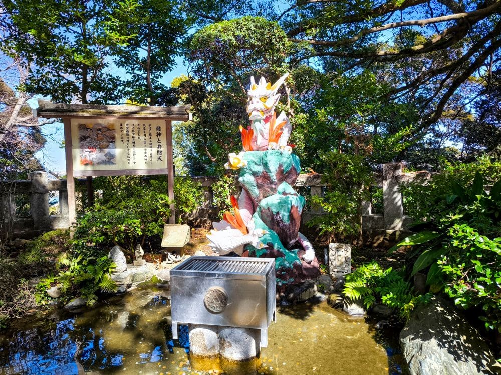 Eine farbenfrohe Drachenstatue sitzt auf einem felsigen Sockel, umgeben von üppigem Grün im Garten des Hetsunomiya-Schreins. Daneben steht ein Reinigungsbecken aus Metall. Das Sonnenlicht dringt durch die Bäume und erhellt diese ruhige Szene.