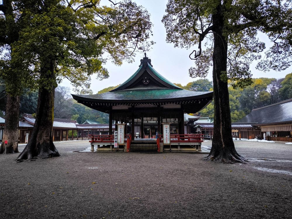 Ein traditioneller Hikawa-Schrein mit grünem Dach steht an einem bewölkten Tag zwischen zwei großen Bäumen, umgeben von Holzgebäuden und Kies.