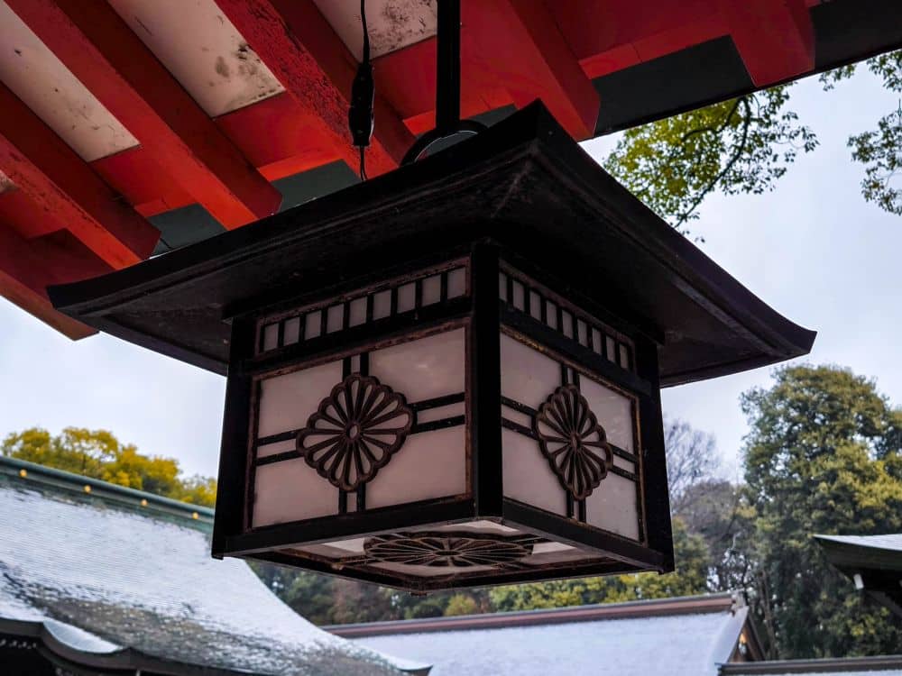 Eine traditionelle japanische Laterne mit dekorativen Blumenmustern hängt unter einem rot-weißen Dach in Hikawa, mit Bäumen und Ziegeldächern im Hintergrund.