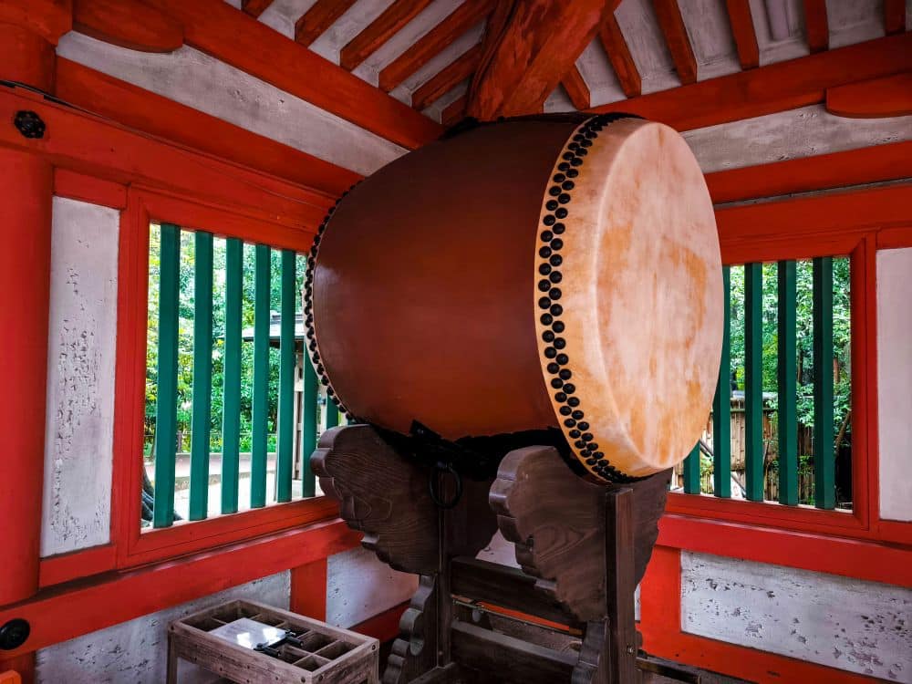 Eine große traditionelle japanische Taiko-Trommel steht in einem Innenraum von Hikawa auf einem Holzständer, umgeben von roten und weißen Wänden mit grünen Holzfenstern. Durch die Fenster ist üppiges Grün zu sehen.