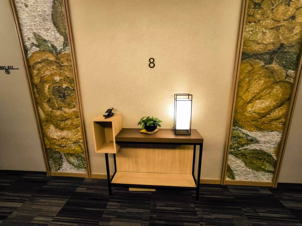Ein kleiner Tobu-Tisch mit einer Topfpflanze, einer Lampe und einer dekorativen Kiste steht an einer Wand mit der Nummer 8, flankiert von großen, blumengemusterten Paneelen und dunklem Teppichboden.