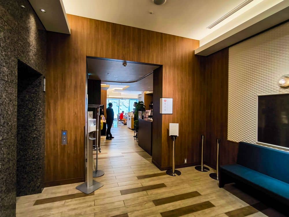Eine moderne Tobu-Hotelhalle mit holzgetäfelten Wänden, einer blauen Bank auf der rechten Seite und einem Flur, der zu einem hell erleuchteten Bereich mit Menschen und großen Fenstern im Hintergrund führt.