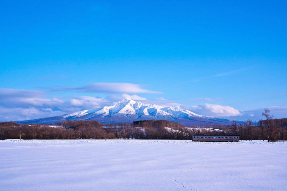 Ein schneebedecktes Feld in Hokkaido erstreckt sich in Richtung eines entfernten Zuges und blattloser Bäume, während sich im Hintergrund ein majestätischer, schneebedeckter Berg unter einem strahlend blauen Himmel erhebt.