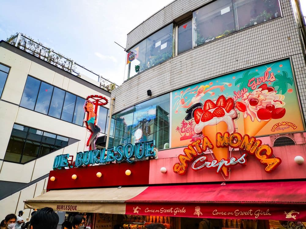 Ein farbenfrohes Schaufenster mit einem "Santa Monica Crepes"-Schild und einer Neon-Burlesque-Tänzerin auf dem Dach. Dieses Harajuku-Lokal in der Takeshita Street ist ein Anziehungspunkt für süße Abenteuer unter der Sonne, umgeben von bunten Plakaten und lebhaften Menschen.