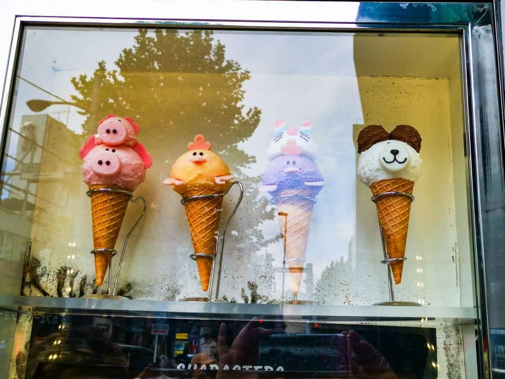 Vier Eistüten mit Kugeln in Form von niedlichen Tiergesichtern (zwei Schweine, ein Hase und ein Hund) sind in einer Glasvitrine in der Takeshita Street in Harajuku ausgestellt. Im Hintergrund sind Spiegelungen von Bäumen und Gebäuden zu sehen.