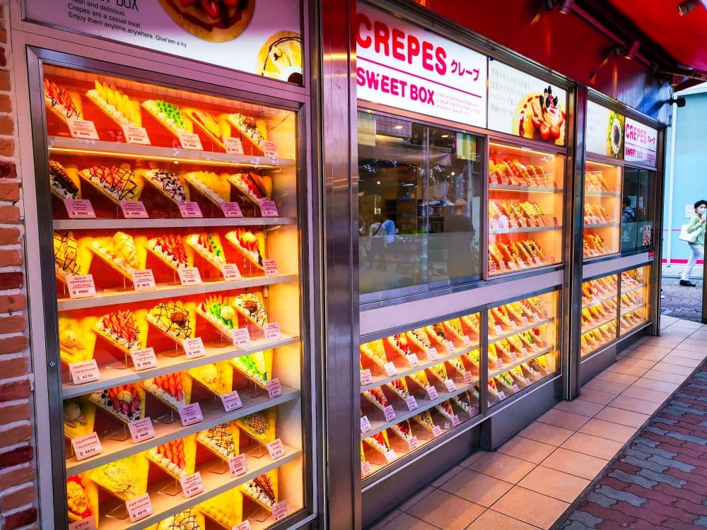 In einem hell erleuchteten Laden in der Takeshita Street in Harajuku sind Dutzende bunter Plastik-Crepes mit verschiedenen Belägen aufgereiht, die jeweils mit einem Preisschild versehen sind. Auf dem Schild des Ladens steht "Crepes Sweet Box" - perfekt für Ihr nächstes Abenteuer.