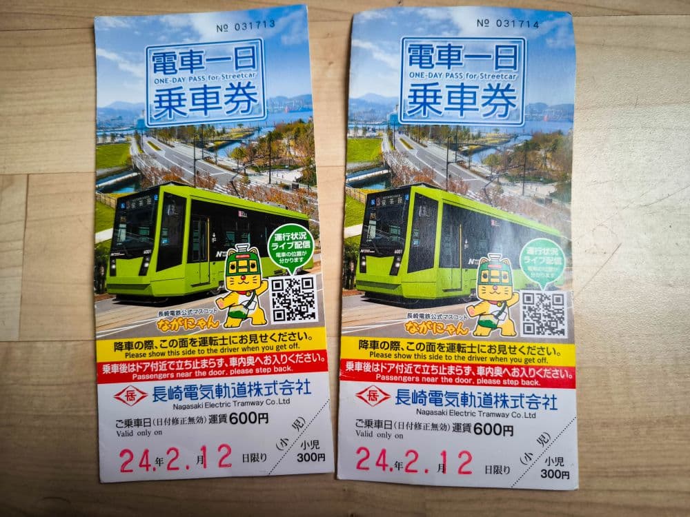 Zwei Nagasaki-Straßenbahn-Tageskarten mit gelb-grünen Nagasaki-Straßenbahnen, die auf den Fahrkarten abgebildet sind, japanischem und englischem Text, einer Cartoon-Straßenbahn-Figur, QR-Code und dem Datum 24.2.12 in Rot am unteren Rand. Die Fahrkarten sind auf einer Holzoberfläche.