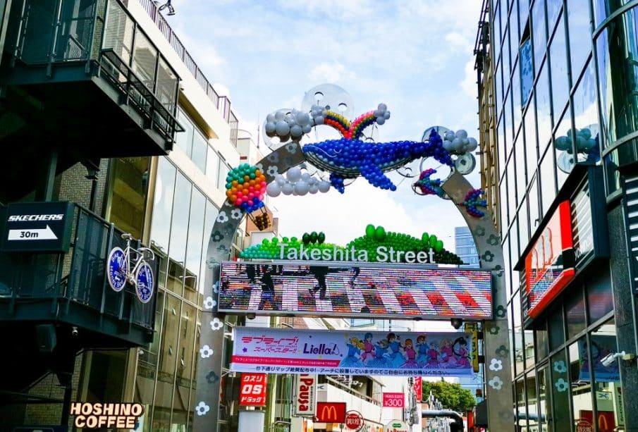 Der Eingangsbogen zur Takeshita Street in Harajuku, Tokio, ist mit bunten Luftballons in Form eines Wals und Wolken geschmückt, umgeben von modernen Gebäuden, Geschäften und sichtbaren Schildern wie McDonald's und Hoshino Coffee.
