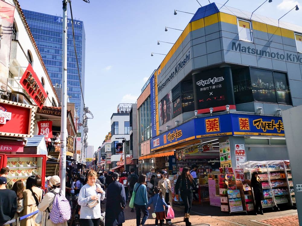 Eine belebte Straßenszene in Japan mit vielen Menschen, die an den Geschäften in der Takeshita Street in Harajuku vorbeilaufen, darunter eine farbenfrohe Apotheke von Matsumoto Kiyoshi. An den Gebäuden sind Schilder angebracht, und der Himmel ist klar und sonnig, was das Gefühl von Abenteuer noch verstärkt.