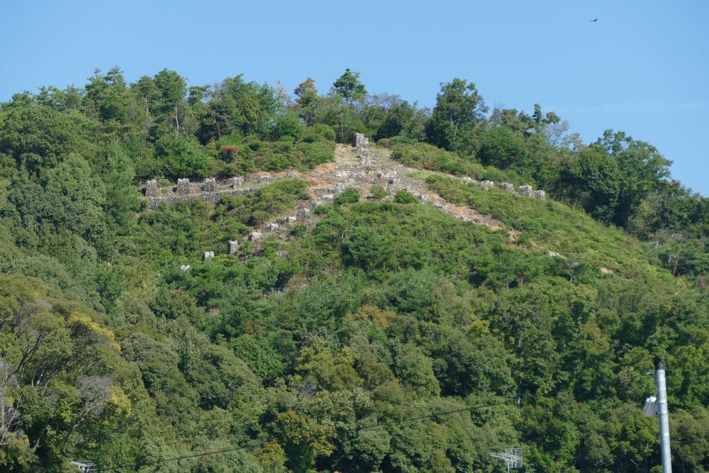 Auf einem grünen Hügel mit spärlichen Bäumen sind große Steinstrukturen in Form eines riesigen X angeordnet, die an das Daimonji-Festival erinnern. Der Himmel ist klar und blau, und am Fuße des Hügels steht ein weißer Pfahl.