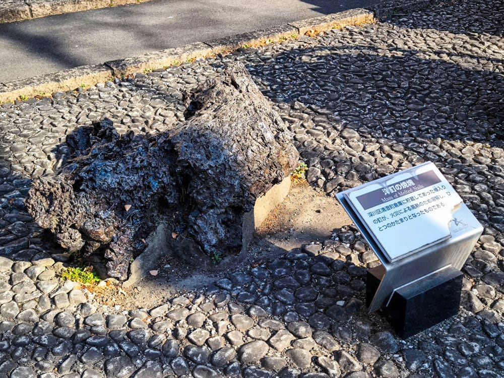 Ein großer, dunkler, unregelmäßig geformter Felsen liegt auf einem Kopfsteinpflaster neben einem Schild mit japanischem Text in einem Außenbereich, in dem das Sonnenlicht Schatten wirft, und erinnert an das Große Kanto-Erdbeben und seinen Platz in der Geschichte. Great Kanto Earthquake Memorial Museum