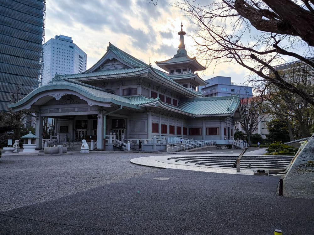 Traditioneller japanischer Tempel mit grünen Ziegeldächern und verschnörkelten Details, der nach dem großen Kanto-Erdbeben unverwüstlich blieb, vor dem Hintergrund moderner Stadtgebäude in der Abenddämmerung. Die gepflasterten Gehwege und Bäume unterstreichen die historische Bedeutung des Ortes. Great Kanto Earthquake Memorial Museum