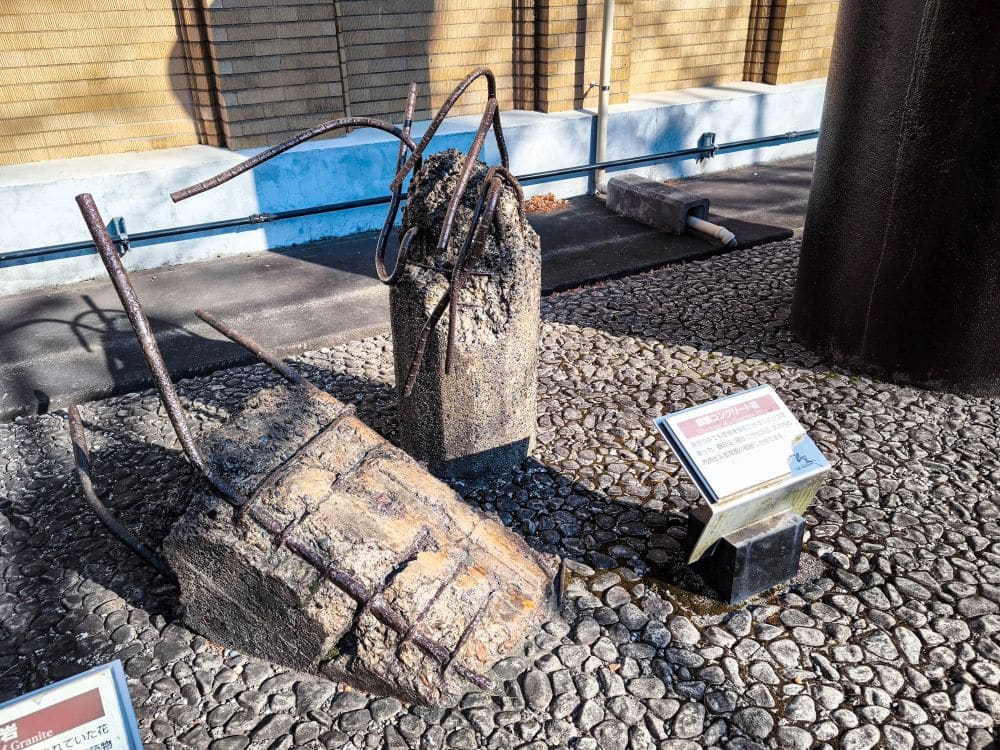 Zerbrochene Betonblöcke mit freiliegenden, verdrehten Bewehrungsstäben sind im Freien auf einem Kieselstein ausgestellt, begleitet von zwei Informationstafeln, die die Geschichte des Großen Kanto-Erdbebens in diesem Gedenkmuseum beschreiben. Im Hintergrund verlaufen eine gelbe Ziegelwand und Rohre. Earthquake